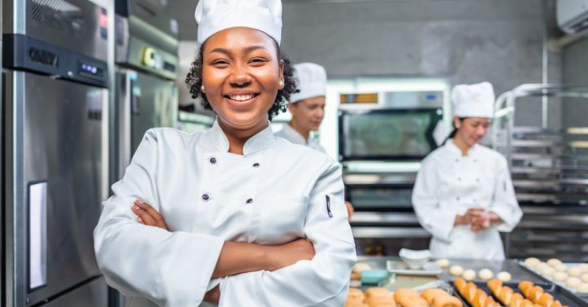 A chef smiles widely as she manages a commercial kitchen in which others are baking pastry.