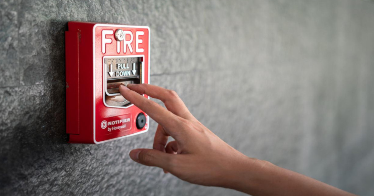 A close-up image of an outstretched hand reaching for a red fire alarm mounted on a grey wall.