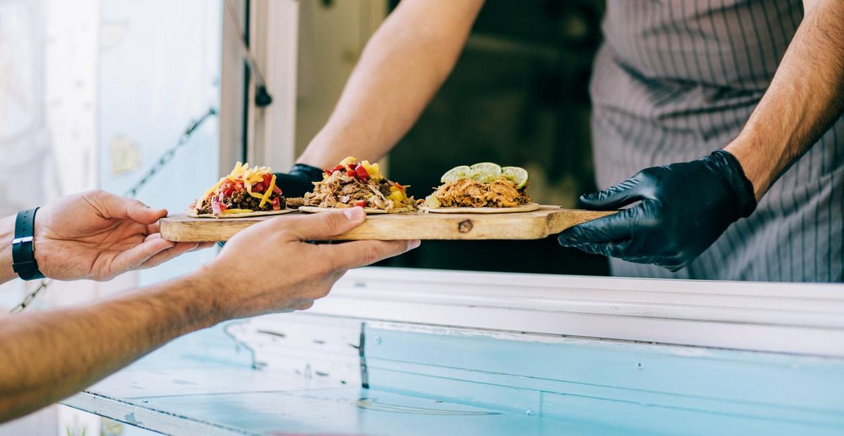 A food truck operator passes a tray of tacos to a happy customer.